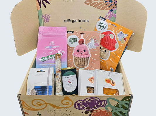 Open box with various items including a mushroom and cupcake DIY crochet kit, dried fruits and nuts, bath soak, delicious matcha sencha tea, vibrant silicone tea steeper, and small batch peanut butter inspired coffee beans, brought to you by Grounded Living