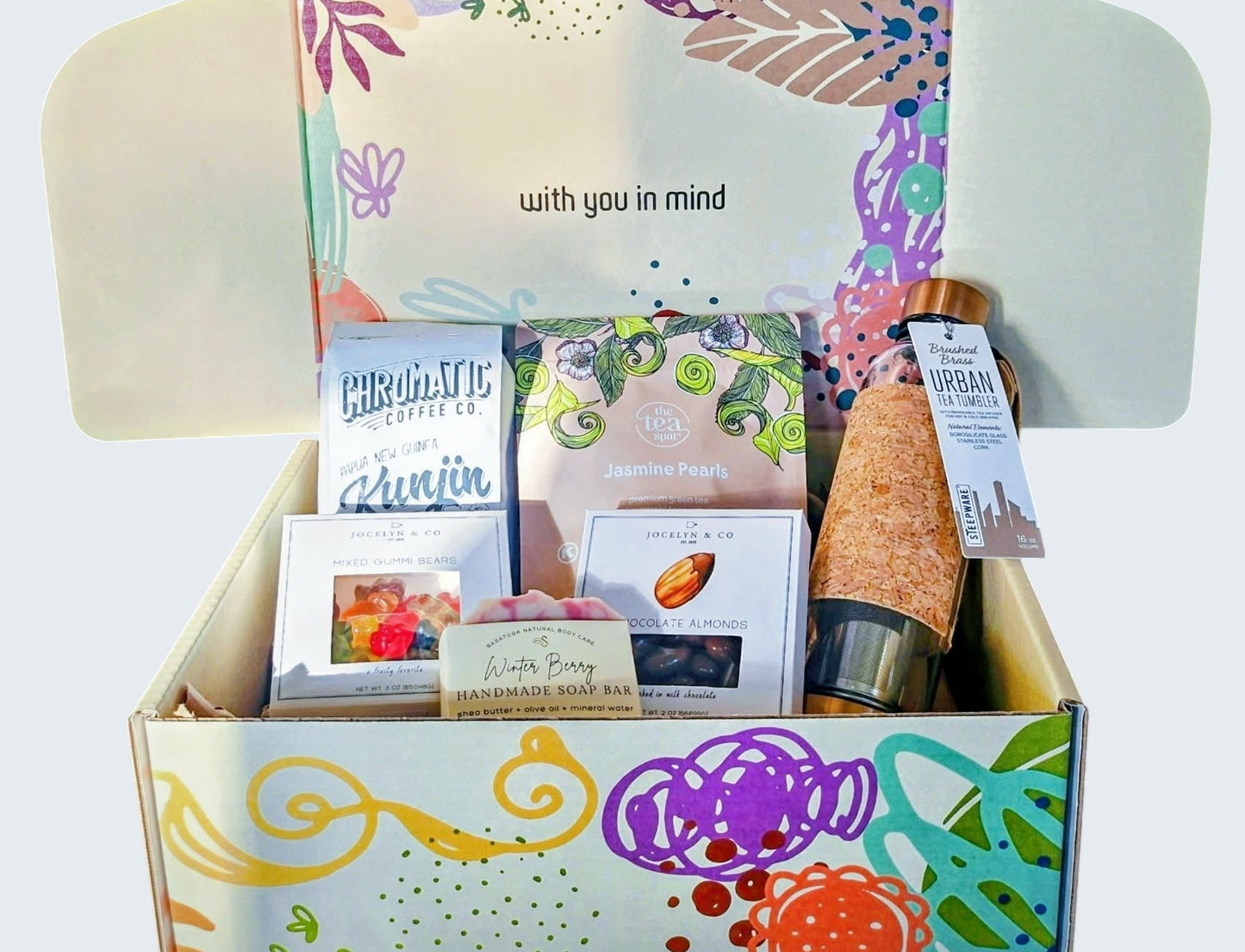 Colorful box featuring beautiful and functional tea tumbler with bronze finishings made of quality tempered glass. Jasmine pearls, mixed gummies, milk chocolate almonds, fragrant soap, and quality small batch coffee from award-winning Chromatic Coffee based in San Jose, CA