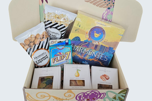 Game Night Box including Harmonies and Flip7 board games, gourmet caramel and white cheddar popcorns, mixed gummies, dried pears, and candied pecans for a fun night in.
